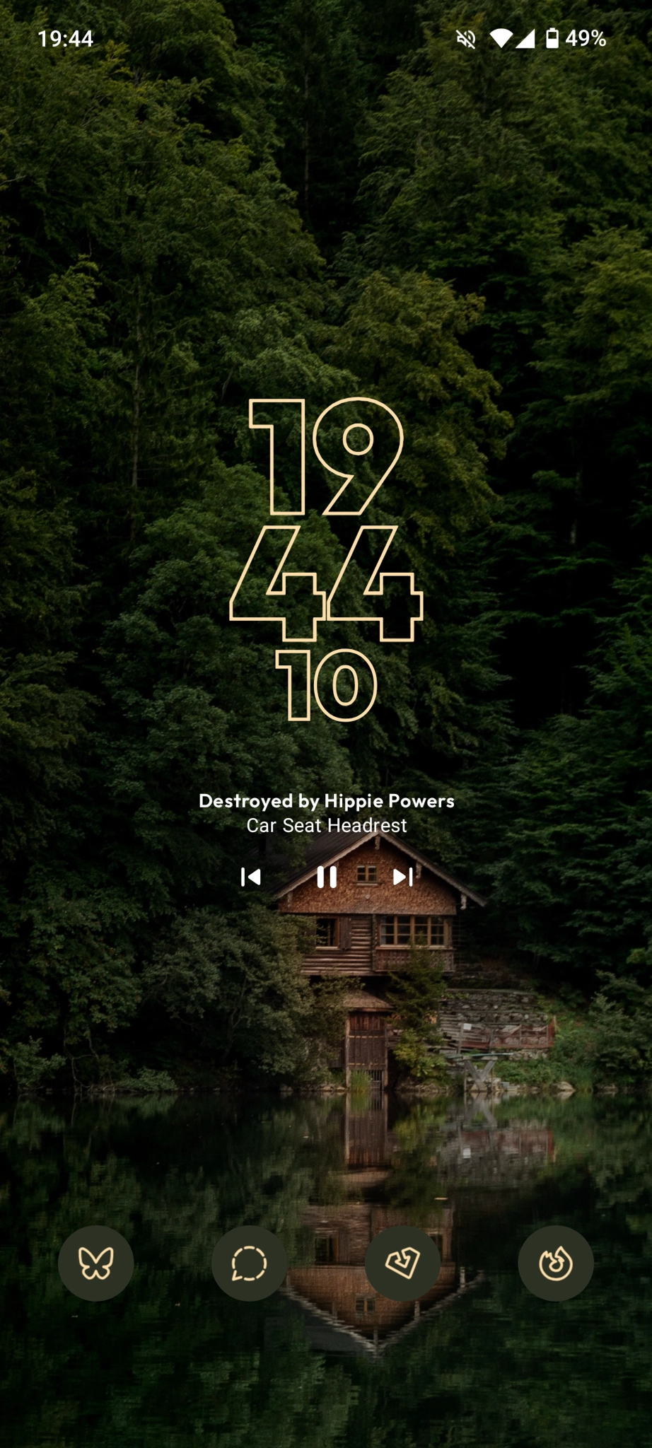 Screenshot of an Android phone running the Kvaesito launcher. The wallpaper is a dark, slightly blurred image of a dilapidated wooden building surrounded by dense, green foliage. The time '19:44' is displayed in a distinctive, slightly distorted font overlaid on the image.  The bottom of the screen shows app icons for Bluesky, Signal, Obtainium, and Firefox