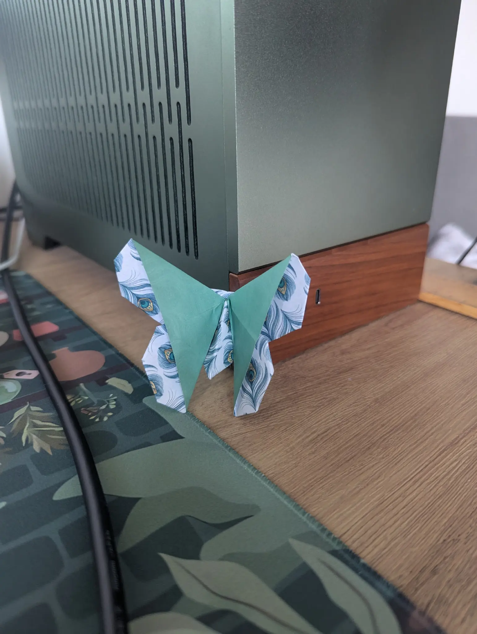 origami butterfly with green wings and peacock feather wing tips and body rested on a computer 