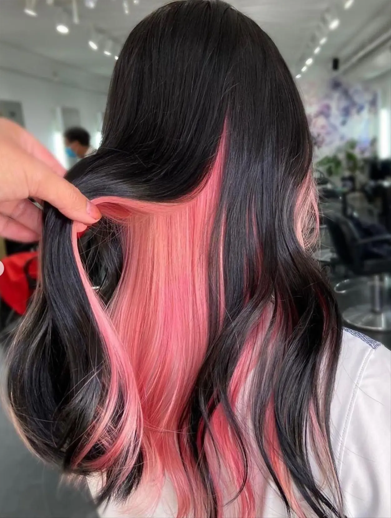 back of a woman's head at a salon where someone is picking up her hair to reveal that under her natural dark brownish hair is a very light pink